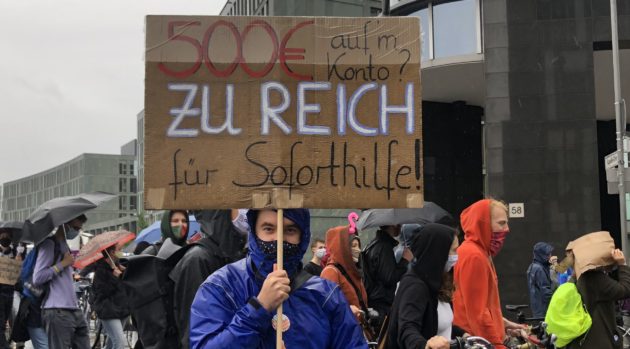 Studierender Demonstrant hält ein Schild mit der Aufschrift „500€ aufm Konto? Zu reich für Soforthilfe?“.