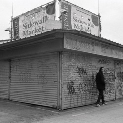 Cover von EVIDENCE - Jim Dean; Schwarzweiß-Foto von einer heruntergekommenen Straßenecke, heruntergelassene Markisen, darüber der Schriftzug "Sidewall Market", eine Person läuft vorbei