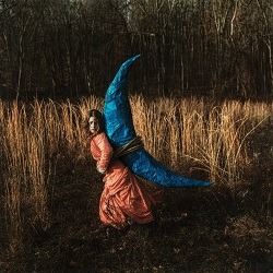 Albumcover von HOLLY MIRANDA – Exquisite; Eine weiße Frau in einem pfirsichfarbenen Kleid auf einem Feld; sie trägt eine blaue Mondsichel auf dem Rücken