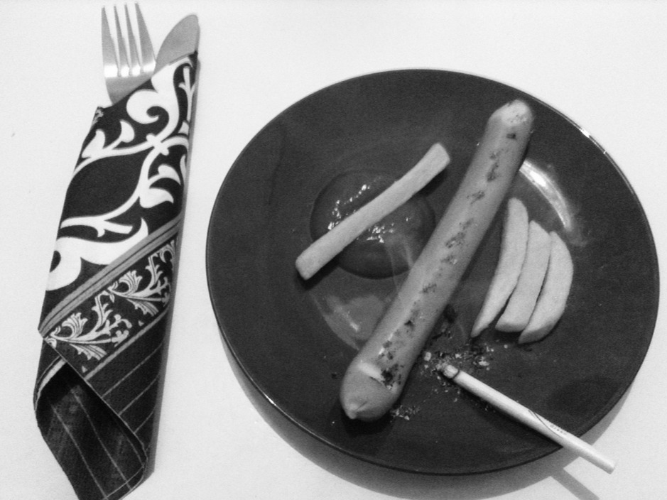 Schwarz-Weiß-Bild mit einem Teller auf dem ein Würstchen, Pommes, Ketchup und eine brennende Zigarette liegen.