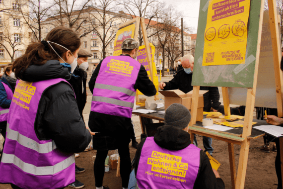 Aktivist:innen in violetten Warnwesten mit dem Aufdruck "Deutsche Wohnen & Co enteignen" stehen vor einem Infostand, an dem Unterschriften gesammelt werden